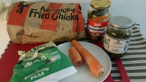 A box of  fried chicken from a well-known chain. A bag of peas, some raw carrot, a jar of mixed pickled veg, and a jar of pickled jalapenos.