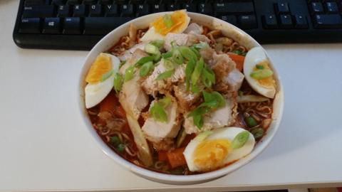 A bowl of instant noodles with added vegetables and chicken pieces, garnished with a quartered boiled egg and chopped spring onion.