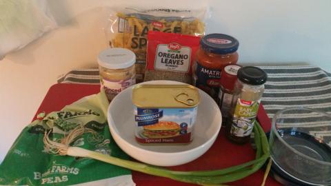 A tin of spam surrounded by seasonings and vegetables, with a packet of spiral pasta and a jar of convenience pasta sauce.