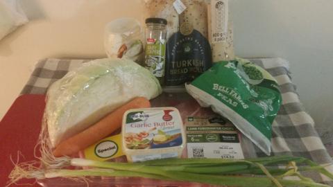 A plastic-wrapped tray of lamb chops, a segment of cabbbage, a carrot, two sprigs of spring onion, peas, a pack of turkish bread rolls, a bottle of aoili and a small tub of garlic butter.