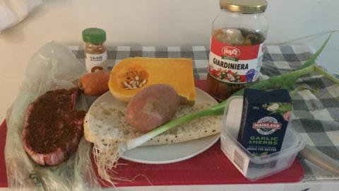 Food preparation area with raw ingredients: seasoning powder, carrot, pumpkin, potato, spring onion, a lamb chop, half a Turkish bread roll, a package of garlic and herb butter.