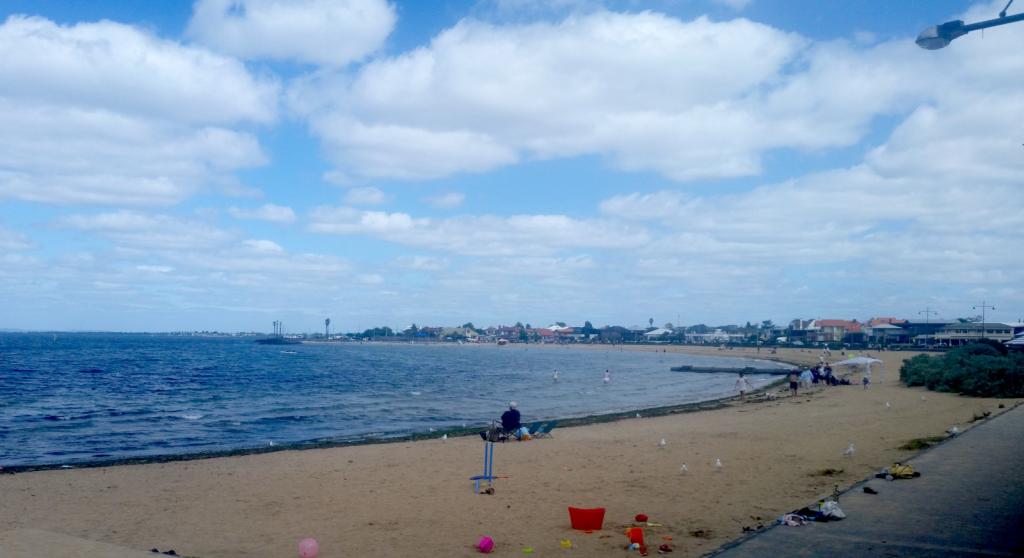 Williamstown Beach. A long, curved stretch of sand. In the foregound, abandoned brightly coloured plastic beach toys, behind them a man in a folding camp chair looks out to sea.