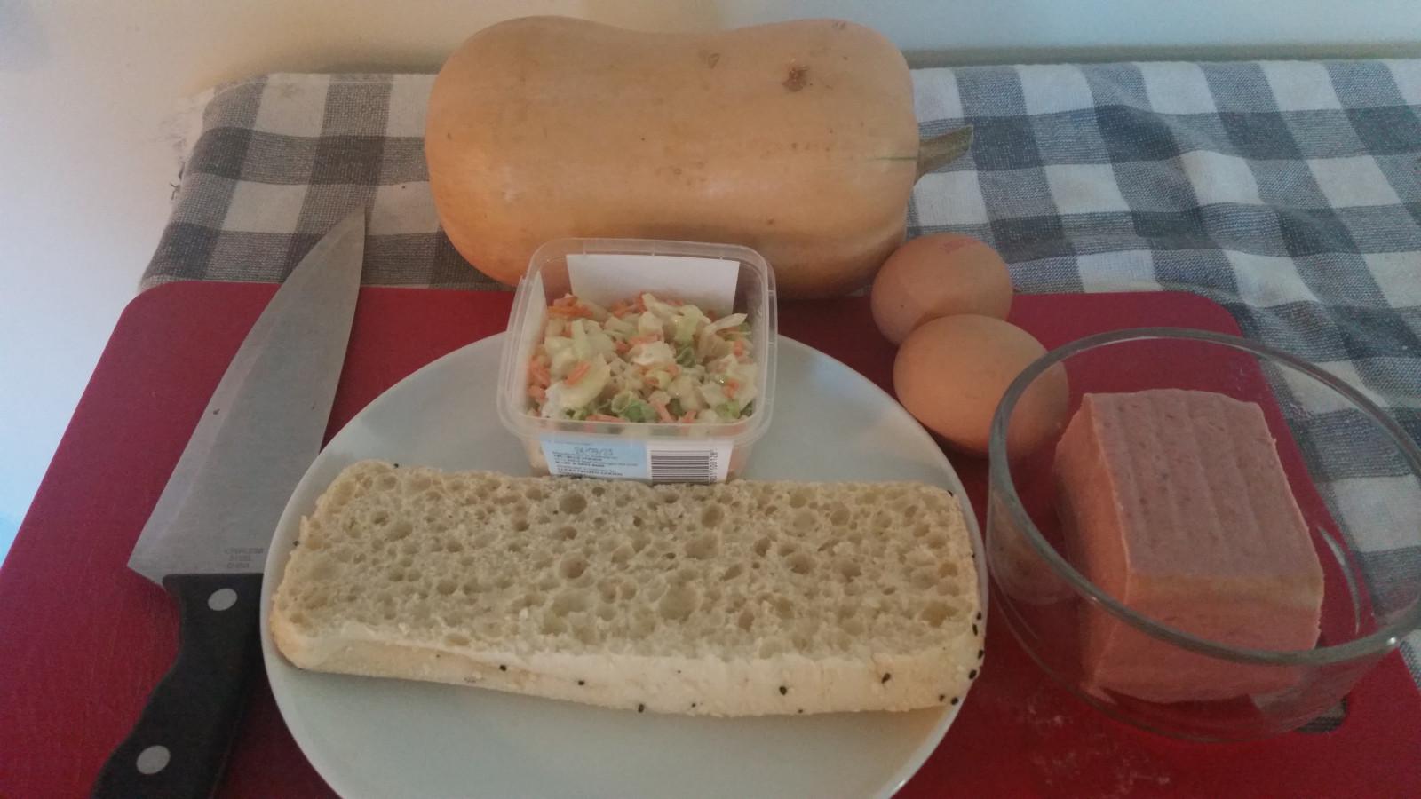 Laid out on a food preparation surface: a butternut pumpkin, a small plastic container of coleslaw, half a Turkish bread roll, two eggs, and a clear bowl containing most of a small tin's worth of ham.