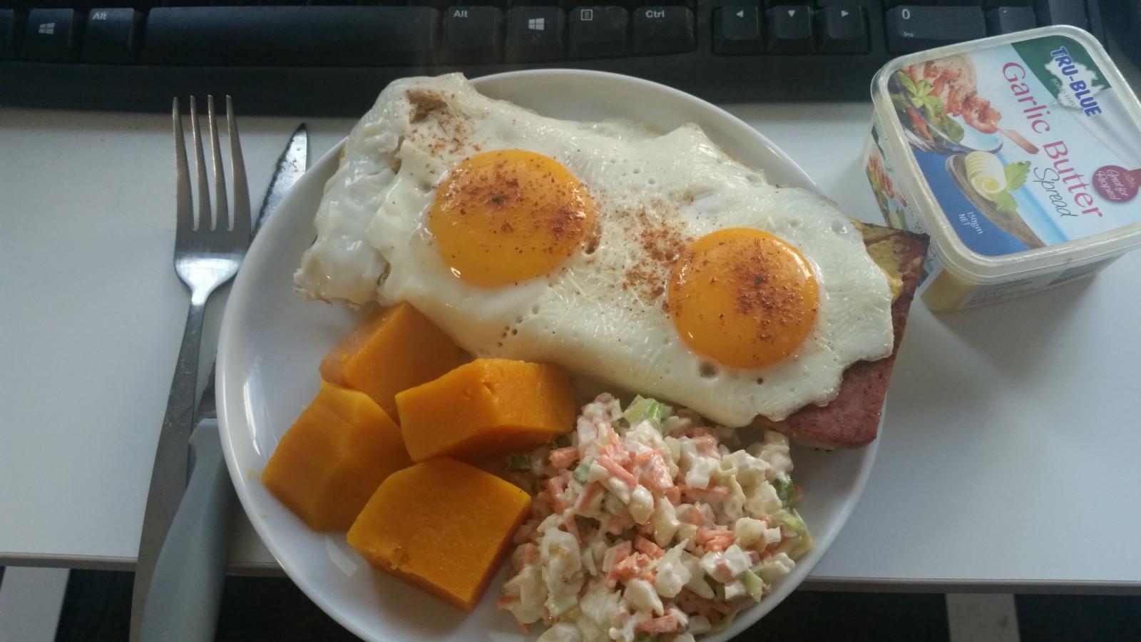 A small plate with two fried eggs on top of some slices of fried ham on toasted Turkish bread, with cubes of boiled pumpkin, and a portion of coleslaw.