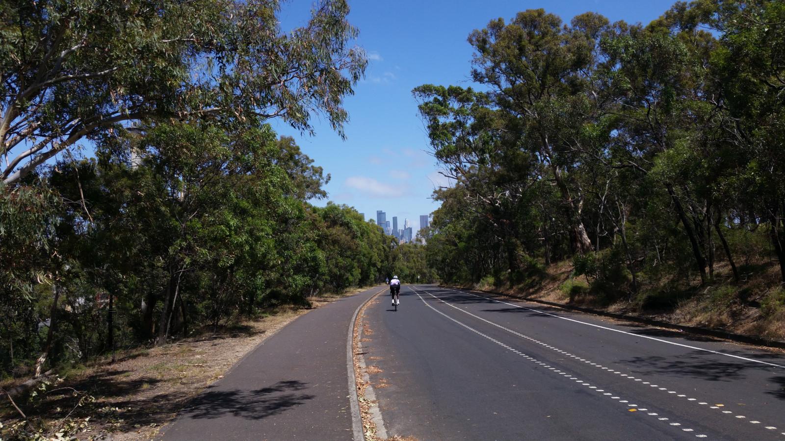 A long stretch of empty road with trees either side. The land slopes from high ground to the right, falling off to the left. City skyscrapers can be seen in the distance.