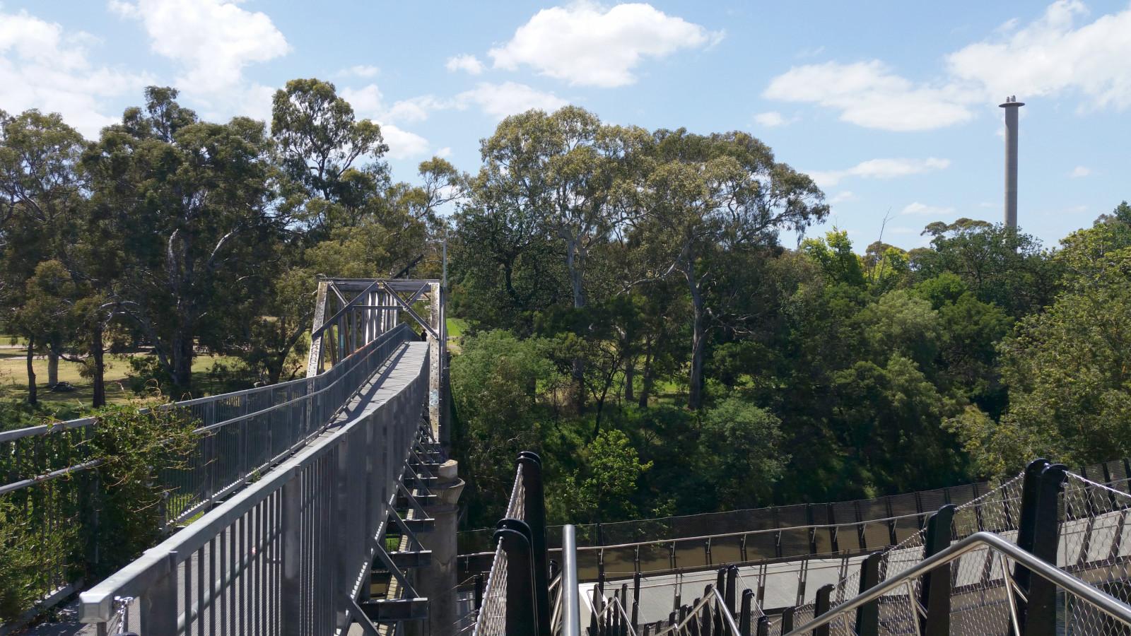 View of the far side of a river. To the left of frame is a bridge across the river. In the foreground at right is a flight of stairs leading down to a ramp which in turn leads down to (out of frame) the river bank.