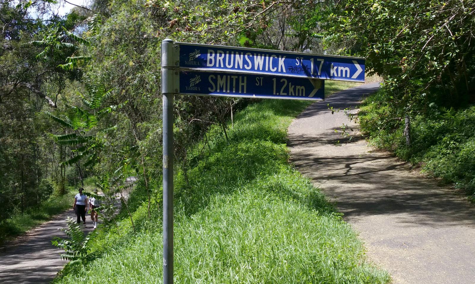 A fork in a pedestrian/cycle trail. To the left a level path, to the right a fairly steeply sloping path and a sign saying "BRUNSWICK STREET 1.7km. SMITH ST 1.2 km."