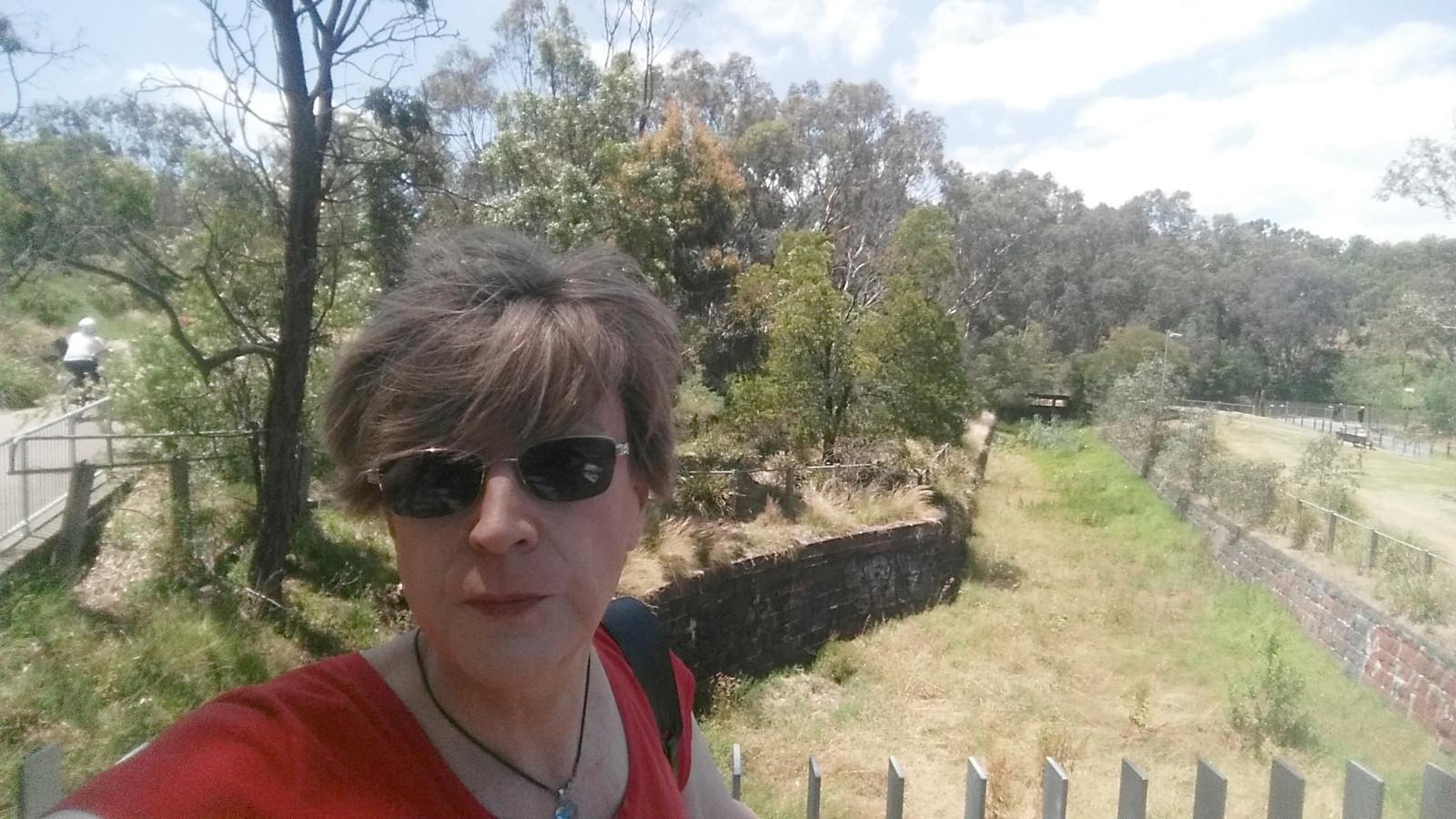 An old lady stands on a viewing platform overlooking a dry, shallow canal overgrown with grass. That is, the canal is overgrown with grass, not the old lady, who is instead wearing dark glasses and a pained expression, as she as at a point in a long walk that is significantly further from invigoration than exhaustion.
