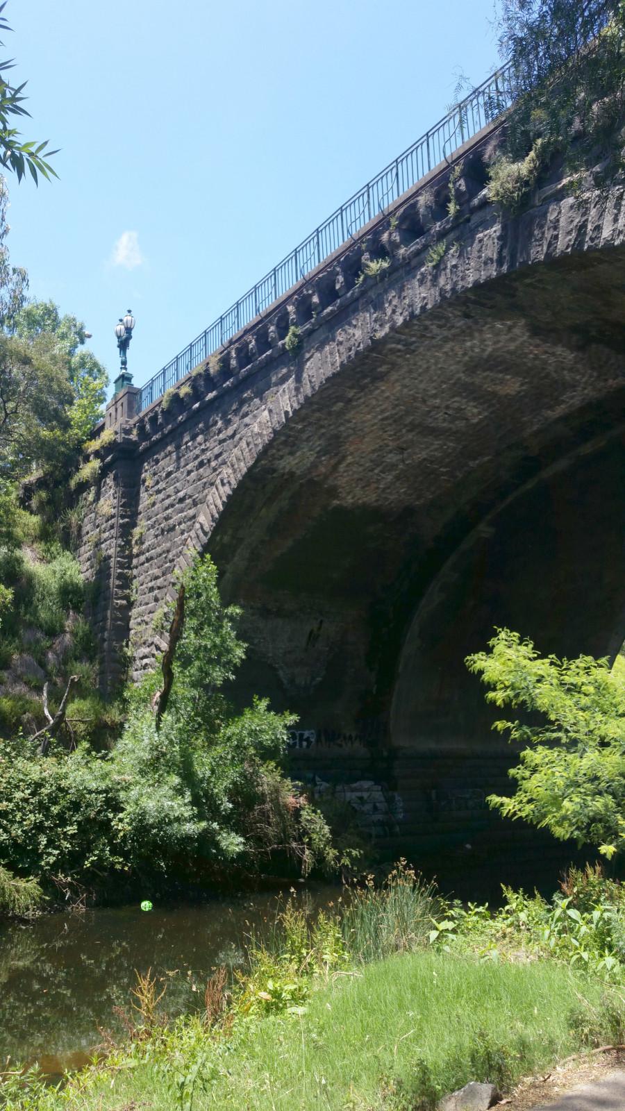 A view of the underside of the Heidelberg Rd bridge over Merri Creek, a blue stone arch decorated with ornate lamps at either end. The creek banks are covered in lush green grasses and shrubs.